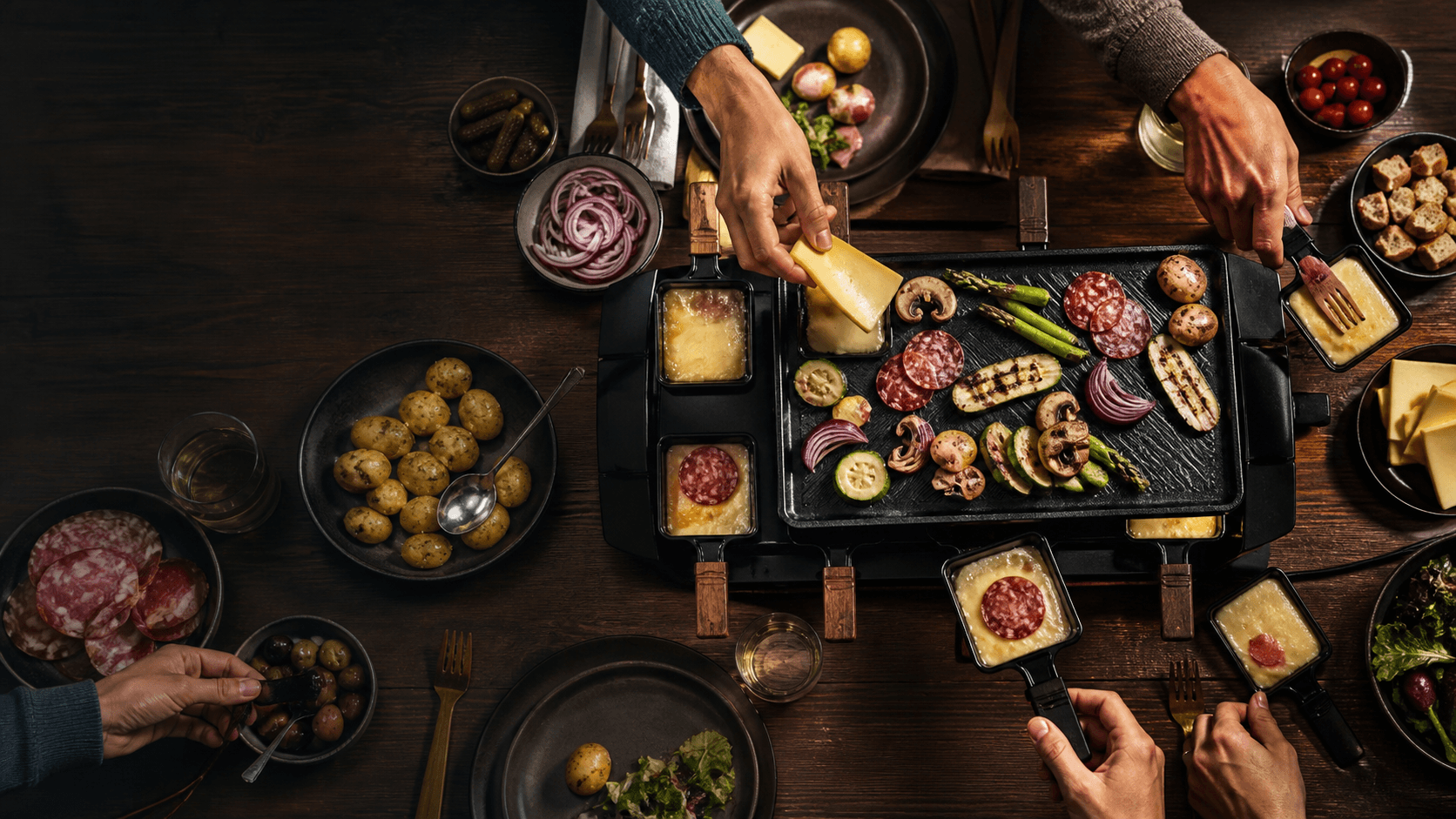 Raclette-Grill mit buntem Gemüse, Kartoffeln, Käse und vielen Händen am Tisch